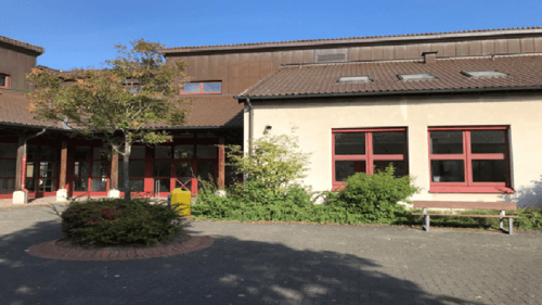 Ein zweistöckiges Gebäude mit einem braunen Dach und rot umrahmten Fenstern steht in einem Innenhof, auf dem ein Baum und eine Bank stehen.