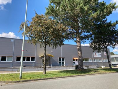 Ein graues, einstöckiges Industriegebäude mit Flachdach und einem Kolping-Schild steht an einer Straße, mit zwei Bäumen und einem blauen Himmel im Hintergrund.