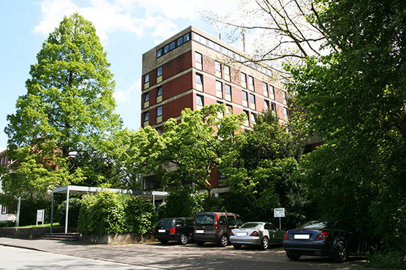 Ein rotes Backsteingebäude mit Flachdach und zahlreichen Fenstern steht inmitten einer Grünanlage, mit einem Parkplatz und Bäumen im Vordergrund.