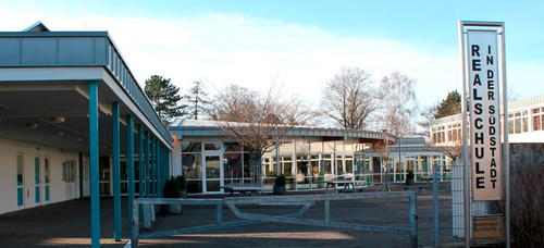 Ein Schulgebäude mit einem teils überdachten Pausengang und einem Schulhof, an dem ein Schild "Realschule in der Südstadt" steht.