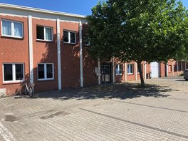 Ein rotes Backsteingebäude mit weißen Streben befindet sich im Hintergrund einen Parkplatzes, auf dem ein Baum und ein Fahrrad in einem Fahrradständer stehen.