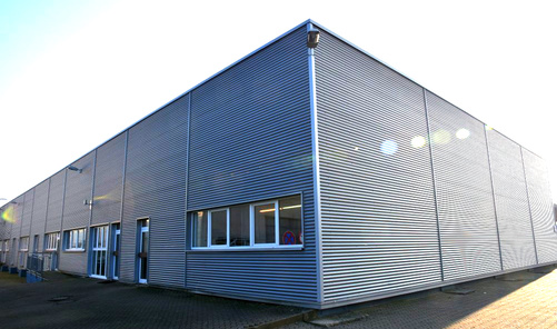 Ein graues, einstöckiges Gebäude mit Flachdach und gewellter Fassade  wird von der Sonne beschienen