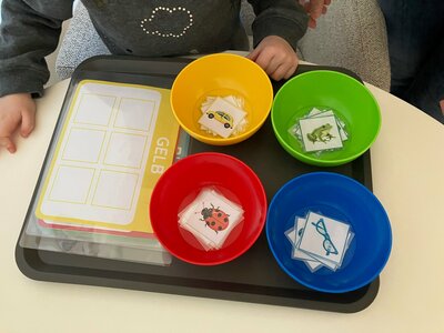 Ein Kind steht vor einem Tablett mit einer gelben, einer roten, einer grünen und einer blauen Schale. In den Schalen liegen Bilder von einem Auto, einem Frosch, einem Marienkäfer und einer Brille. Neben den Schalen liegt eine Aktionskarte.