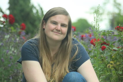 Ein Foto von einer jungen Frau vor einem Blumenbeet mit blauen und roten Blumen.