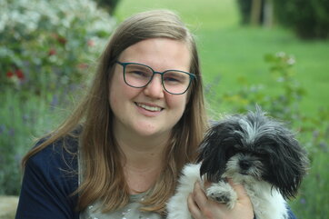 Eine lächelnde Frau hält ihren Shih-Tzu-Zwergspitz-Mix im Arm, während beide in einem üppig-begrünten Garten sitzen.