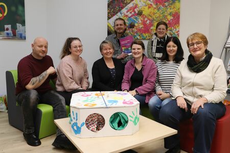 Eine Gruppe von acht Personen gruppiert sich in einer Schulbibliothek um einen Tisch, auf dem eine Fühlbox steht.