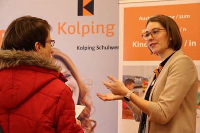 Eine Frau befindet sich bei einer Berufsmesse am Messestand des Kolping Schulwerks im Gespräch mit einer Besucherin und gestikuliert mit den Händen.