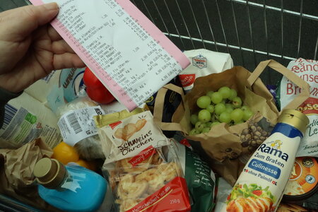 Ein Einkaufswagen ist gefüllt mit zahlreichen Lebensmitteln, darüber wird von einer Hand ein rosa-umrandeter Kassenzettel gehalten.