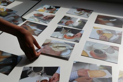 Eine Hand legt ein Foto auf einen Tisch, auf dem eine Vielzahl an Fotografien verteilt liegt, auf denen verschiedene Frühstückszutaten und Utensilien abgebildet sind.