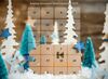 Ein Adventskalender mit weißen und blauen Weihnachtsbäumen, Sternen und einem schneebedeckten Boden wird vor einem rustikalen Holzhintergrund präsentiert.