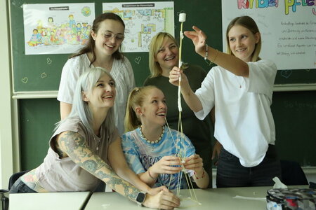 Fünf Schülerinnen arbeiten gemeinsam an einem Projekt, bei dem sie am Pult eines Klassenzimmers aus Spaghetti und Marshmallows einen hohen Turm bauen und dabei viel Spaß haben.