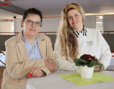 Zwei Frauen, eine in einem beigen Blazer und die andere in einem weißen Pullover, stehen anlässlich eines Fachtags an einem Tisch mit weißer Tischdecke und grüner Serviette und lächeln in die Kamera.