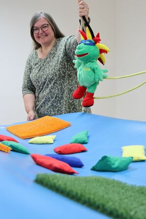 Eine Frau steht lächelnd hinter einer blauen Matte und hält ein grünes Stofftier mit roter und gelber Kopfverzierung hoch in der Hand. Auf der Matte liegen verschiedenfarbige Säckchen.