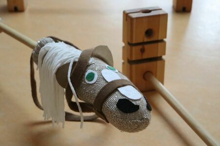 Ein selbstgebasteltes Steckenpferd mit heller Mähne, Zaumzeug und Stock schwebt vor einer Holzstange, die als Hindernis dient.