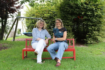 Die beiden Leiterinnen der Kindertageseinrichtung auf der Brede in Brakel sitzen vor einigen Bäumen und einem Spielgerät auf einer roten Bank.