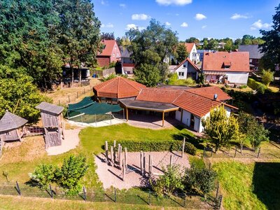 Die Luftaufnahme zeigt die Kita Hüssenbergnest, die umgeben ist von einem weitläufigen, begrünten Außengelände mit Spielplatz.