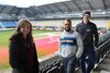 Eine Frau und zwei Männer stehen auf der Zuschauertribüne eines Stadions, das auf dem Hintergrund zu sehen ist. Der grüne Rasen ist durchzogen von roten Flächen.