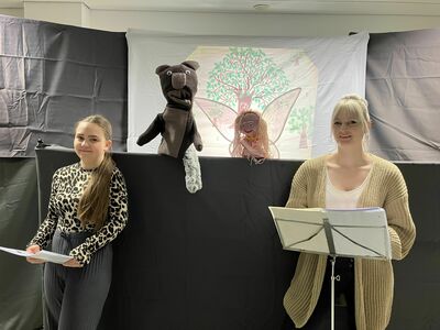 Zwei Frauen, eine mit einem Notenständer, stehen vor einer Puppentheaterkulisse mit einem Eichhörnchen und einer Fee, im Hintergrund hängt das Bild von einem Baum.