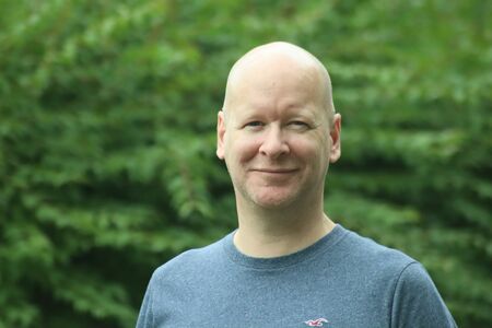 Das Porträt zeigt den stellvertretenden Schulleiter des Kolping-Sozial-Berufskollegs Delbrück vor einer grünen Hecke.