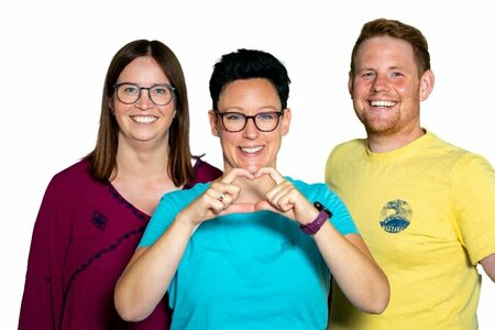 Das Schulleiterteam des Theresia-Gerhardinger-Berufskollegs in Rimbeck präsentiert sich farbenfroh in roter, blauer und gelber Kleidung und lächelt. Die Schulleiterin in der Mitte formt mit ihren Händen ein Herz.