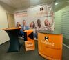 Eine orange-schwarze Theke mit dem Kolping-Schulwerk-Logo, dazu ein orange-schwarzer Stehtisch und orange-schwarze Sitzwürfel stehen vor einer Stellwand mit Kolping-Logos.