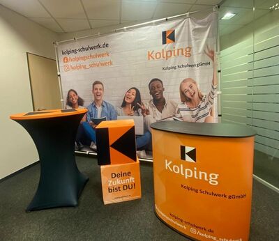 Eine orange-schwarze Theke mit dem Kolping-Schulwerk-Logo, dazu ein orange-schwarzer Stehtisch und orange-schwarze Sitzwürfel stehen vor einer Stellwand mit Kolping-Logos.