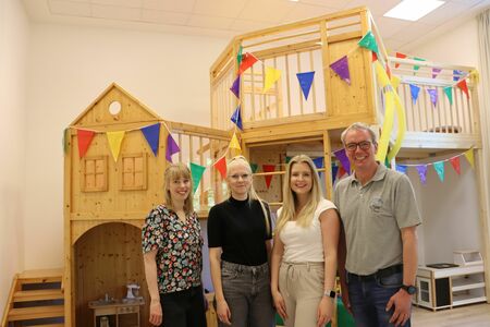 Vier Personen, drei Frauen und ein Mann, stehen vor einem hölzernen Spielhaus mit mehreren Ebenen, das mit bunten Wimpeln geschmückt ist, und lächeln in die Kamera.
