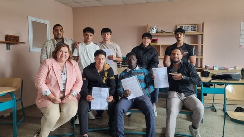 Eine Lehrerin und acht junge Männer sitzen in einem Klassenraum. Ein Teil der Schüler hält stolz Zeugnisse in die Kamera.