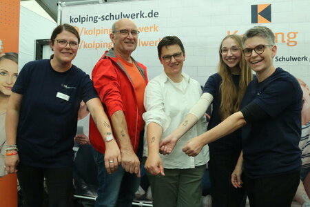 Fünf Personen strecken an einem Messestand ihre Arme in die Kamera, auf denen Glitzer-Tattoos mit dem Schullogo zu sehen sind.