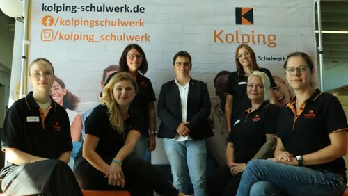 Eine Gruppe von sieben Personen in schwarzen Poloshirts mit orangefarbenen Kolping-Logo posiert vor einer Messestellwand mit dem Kolping-Schulwerk-Logo und Social-Media-Handles.