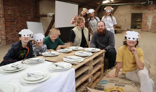 Eine Gruppe Kinder mit Schafsmasken und zwei Erwachsene gruppieren sich um eine Mittagstafel auf dem Heuboden des Kolping Gutshofs