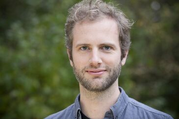 Ein Porträt von einem Mann, der vor einem unscharfen grünen Hintergrund in die Kamera lächelt.