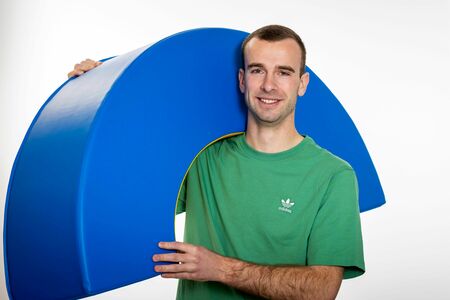 Ein junger Mann im grünen T-Shirt trägt einen blauen, runden Schaumstoff-Baustein über der Schulter.