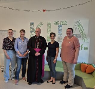 Fünf Personen, darunter ein Weihbischof, stehen im Kindergarten vor einer weißen Wand mit einem Schriftzug.